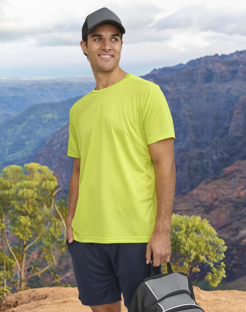 A man wearing a bright yellow short-sleeve performance T-shirt stands against a mountainous backdrop, holding a bag.