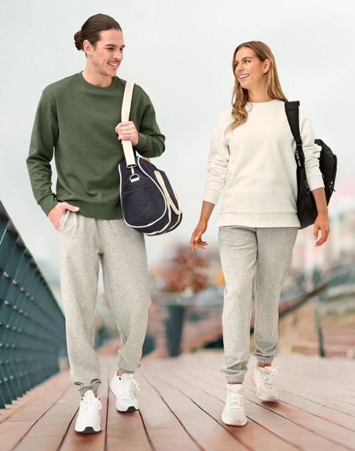 A man in a green sweatshirt and grey sweatpants walks alongside a woman in a white sweatshirt and grey sweatpants, both carrying bags.
