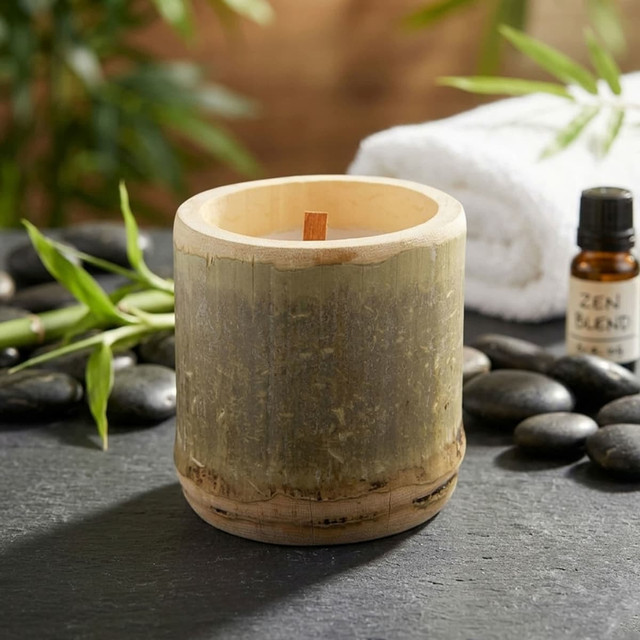 A cylindrical bamboo candle sits on a stone surface, surrounded by pebbles and a towel, with essential oil nearby.