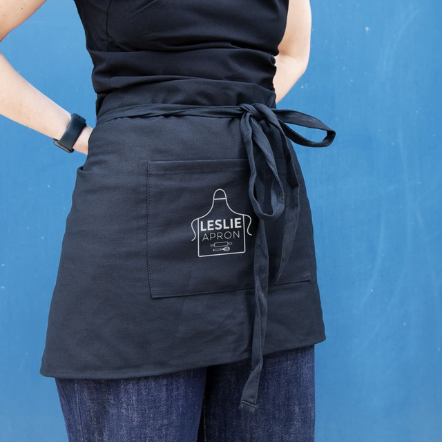 A black waist apron with a front pocket and a tied waist, held by a person against a blue background.