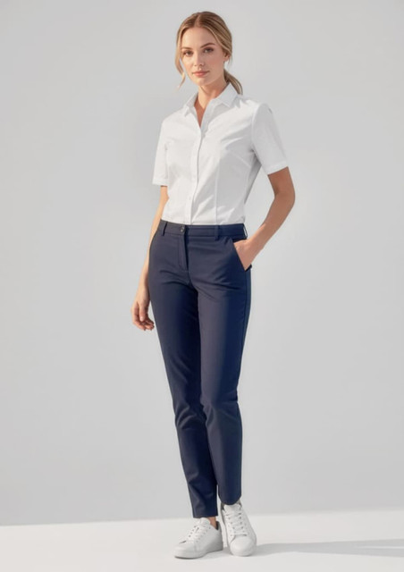 A woman stands wearing navy slim leg pants and a white short-sleeve shirt, with hands in pockets and white shoes.