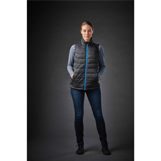 A woman is wearing a black thermal vest with a blue zip, paired with jeans and standing against a dark background.
