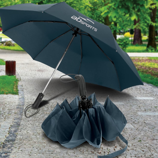 A compact dark teal umbrella with a curved handle, displayed partially open on a path surrounded by greenery.