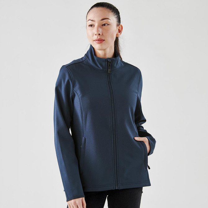 A woman wearing a navy softshell jacket with a high collar and side pockets, featuring a logo on the sleeve.