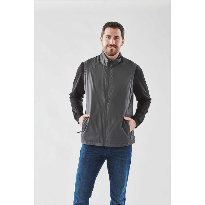 A man wearing a grey vest over a black long-sleeve shirt, posing against a plain background.