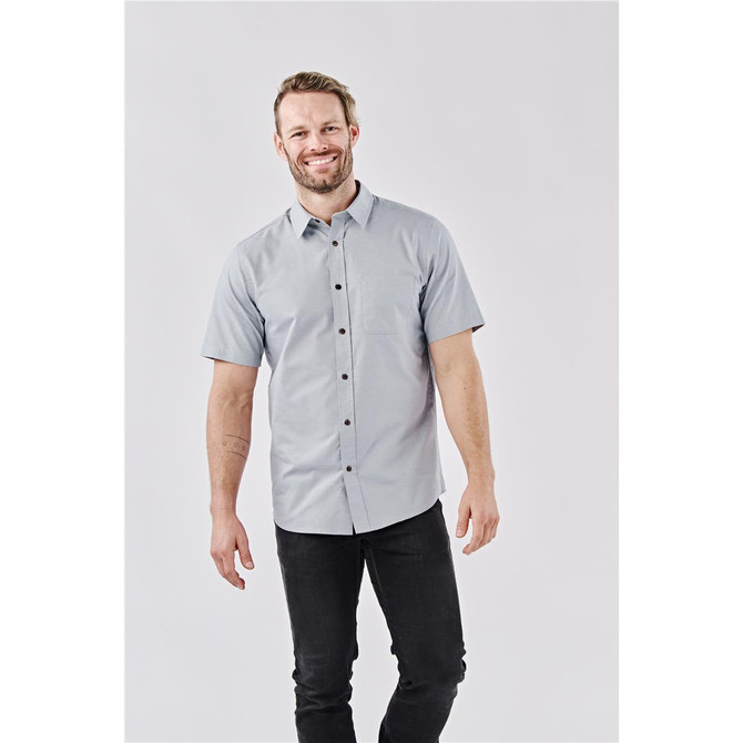 A short-sleeve button-up shirt in light grey, featuring a logo, worn by a smiling male model.