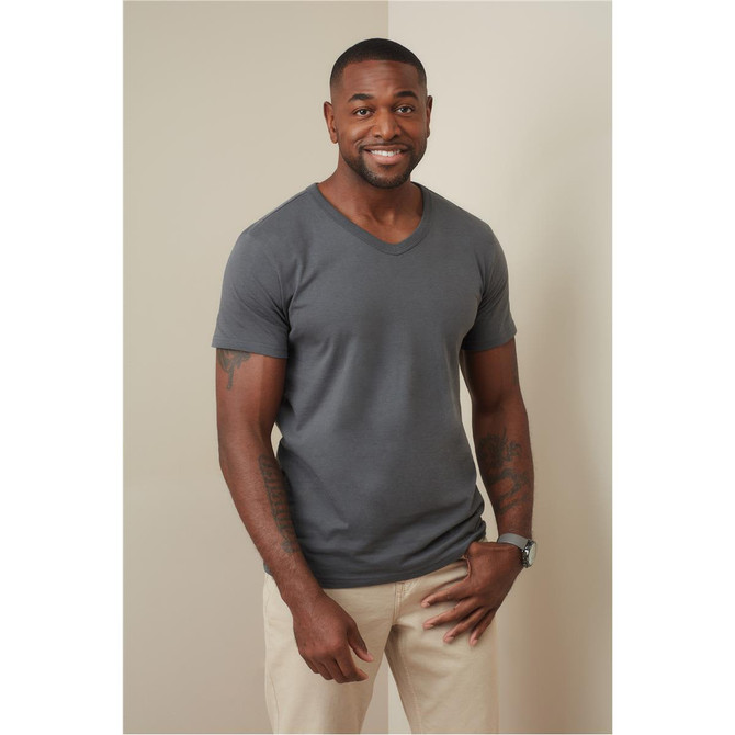 A man in a grey V-neck t-shirt stands against a neutral background, smiling and wearing light-coloured pants.