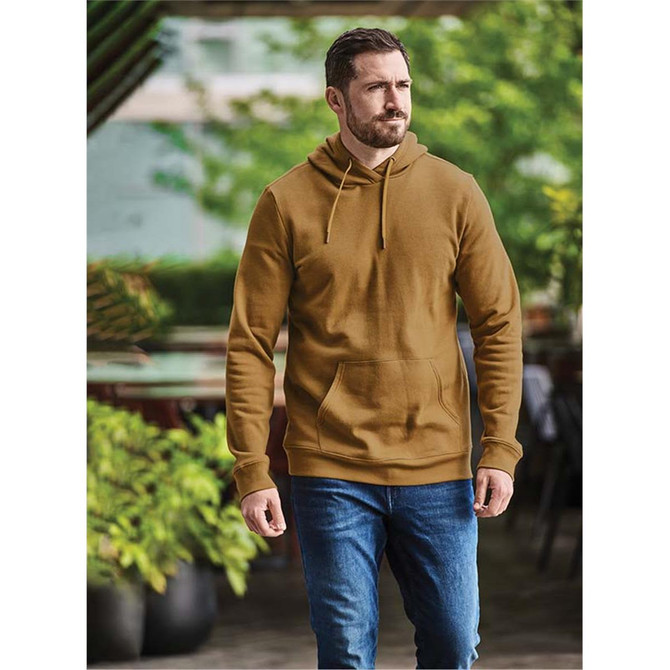 A man wearing a brown fleece pullover hoody, paired with blue jeans, walks outdoors in a leafy setting.