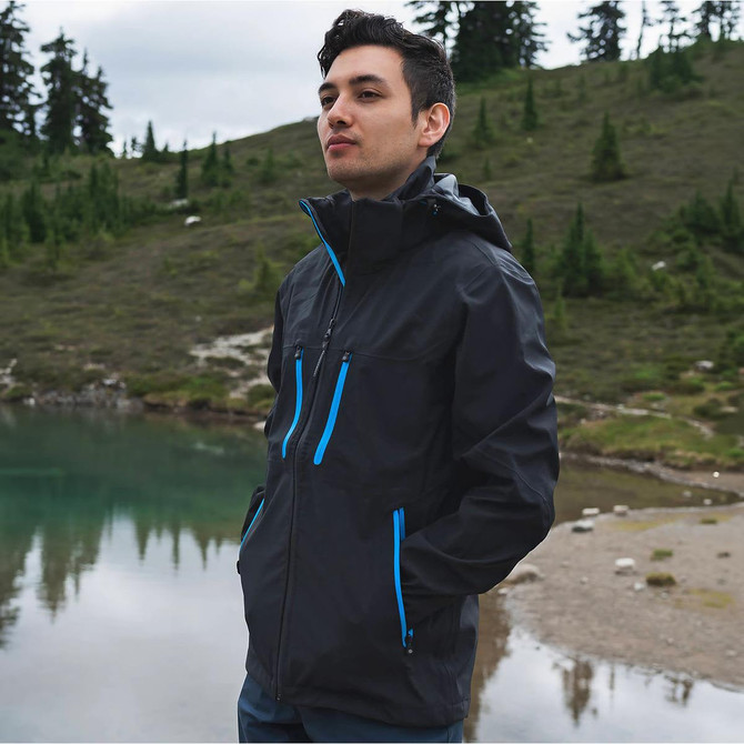 A man stands by a body of water, wearing a black softshell jacket with blue zippers in a natural setting.