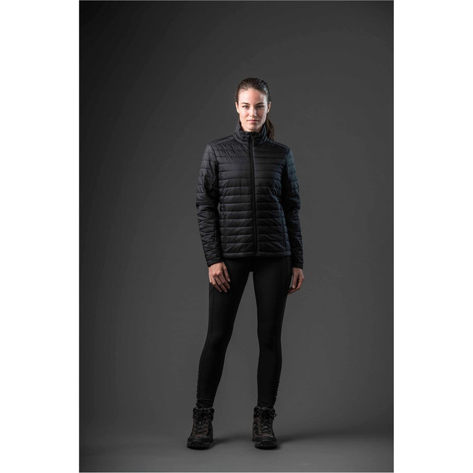 A woman wearing a black padded jacket, black leggings, and boots, standing against a dark background.