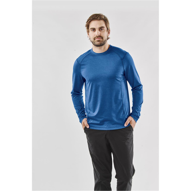 A man wearing a blue long-sleeve crew neck shirt, paired with dark trousers, standing against a plain background.