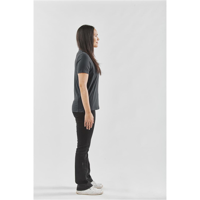 A woman in profile wears a dark polo shirt and black pants, standing against a white background. The shirt has a logo.