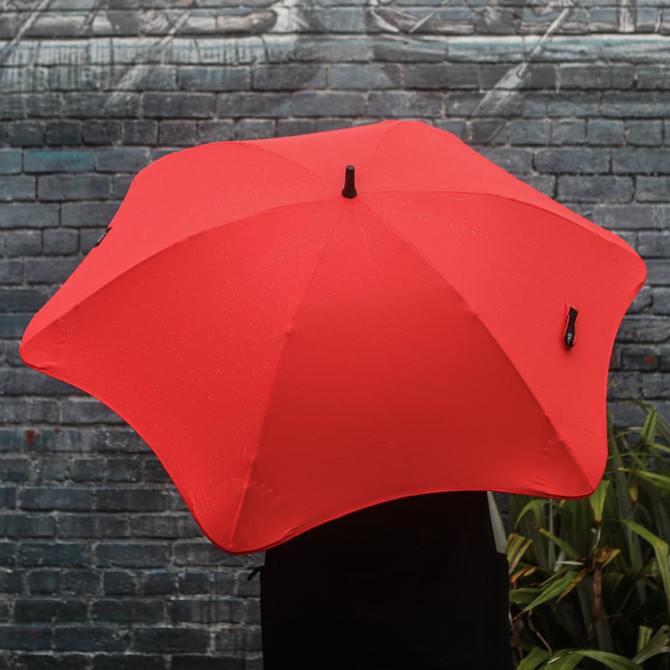 A bright red BLUNT Classic Umbrella with a sleek design and a logo, set against a brick wall background.