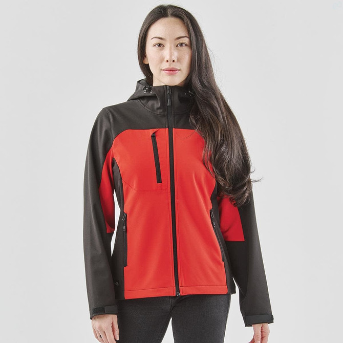 A woman models a red and black hooded softshell jacket, featuring a front zip and side pockets. It has a logo on it.