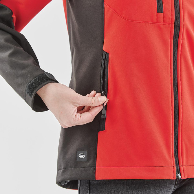A red and black women's softshell jacket featuring zippered pockets and a logo on the lower side.