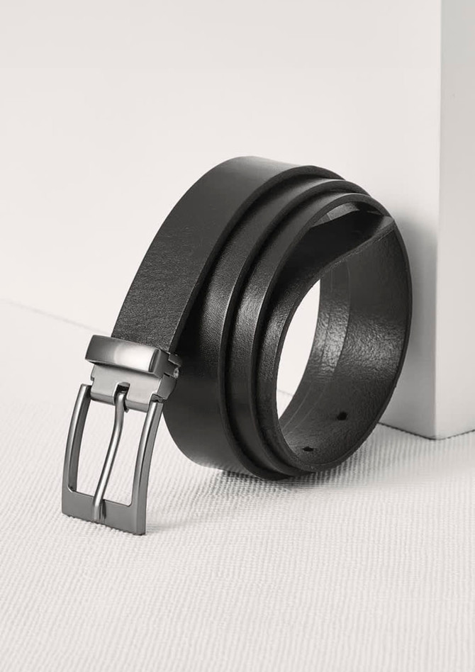 A black leather belt is displayed with a sleek silver buckle, leaning against a white surface.