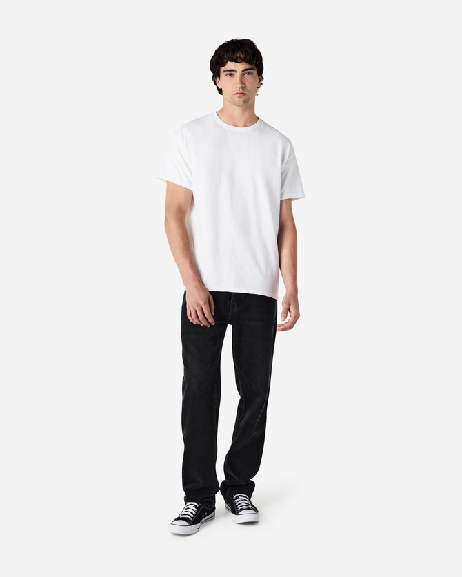 A young man stands wearing a white short sleeve t-shirt and black jeans, with black shoes. The t-shirt has a logo.