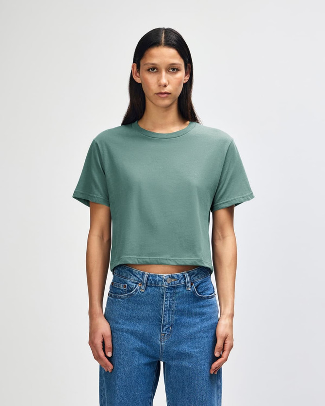 A green women's boxy t-shirt is worn with high-waisted blue jeans, displayed against a neutral background.