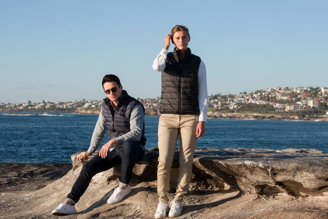A puffer vest in black, worn by two models by the seaside, with a scenic coastal background.