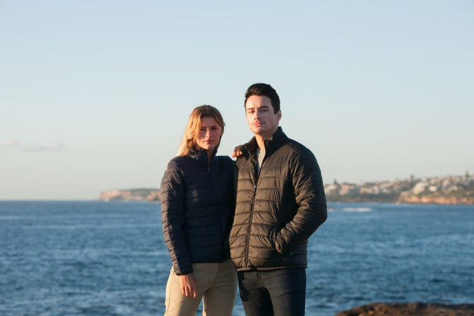 A black puffer jacket worn by a man and a navy puffer jacket worn by a woman against a coastal backdrop.