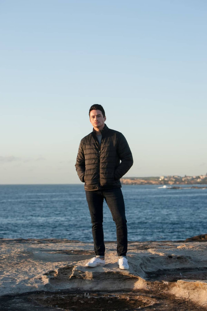 A man stands by the ocean wearing a black puffer jacket, blue jeans, and white shoes, with a coastal background.