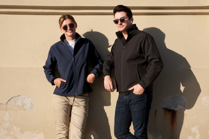 A soft shell jacket in navy worn by a woman and a black jacket worn by a man, both standing against a wall.