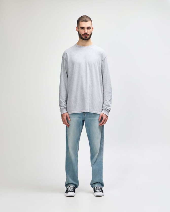 A grey long sleeve T-shirt worn by a man standing against a plain background, paired with light blue jeans.