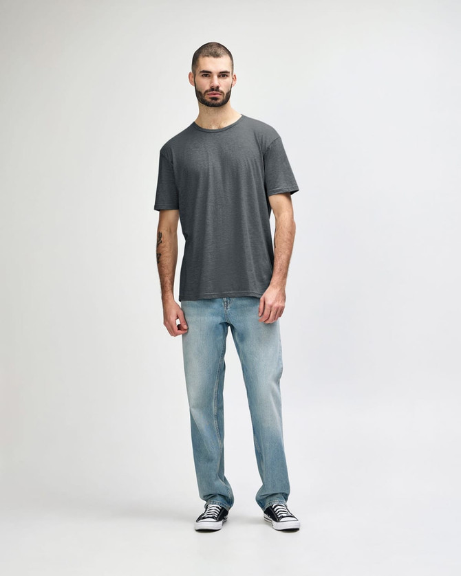 A man stands wearing a grey Gildan Softstyle adult t-shirt paired with light blue jeans and white shoes.