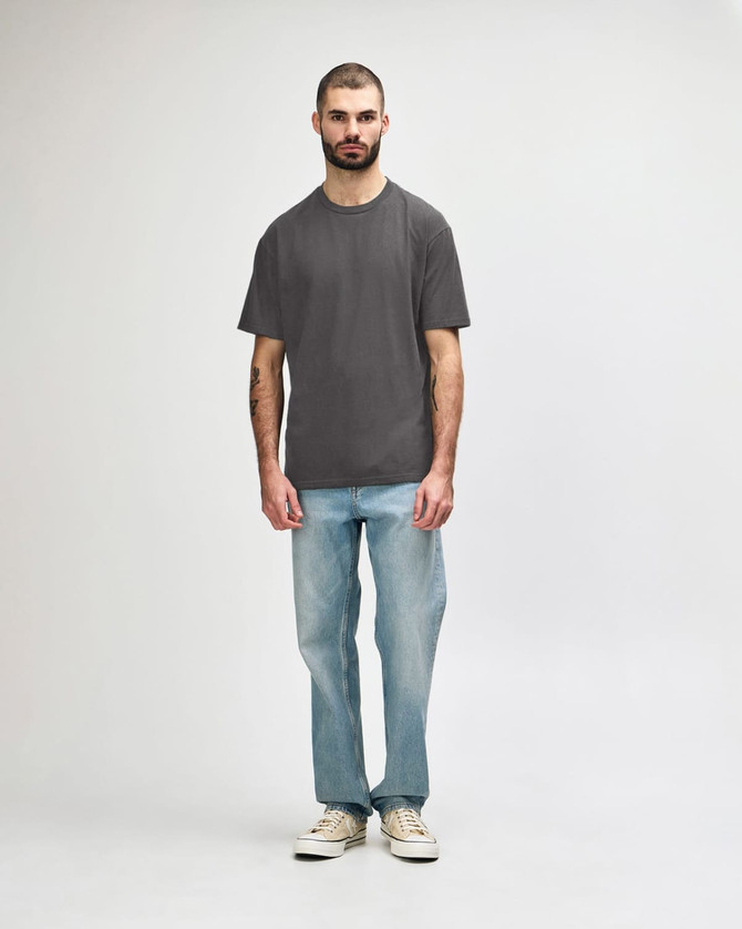 A midweight adult t-shirt in olive green, paired with black jeans and sneakers. The model is walking.