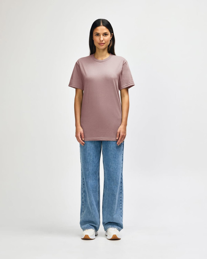 A midweight adult T-shirt in soft pink, worn with black jeans and sneakers. Features a logo.