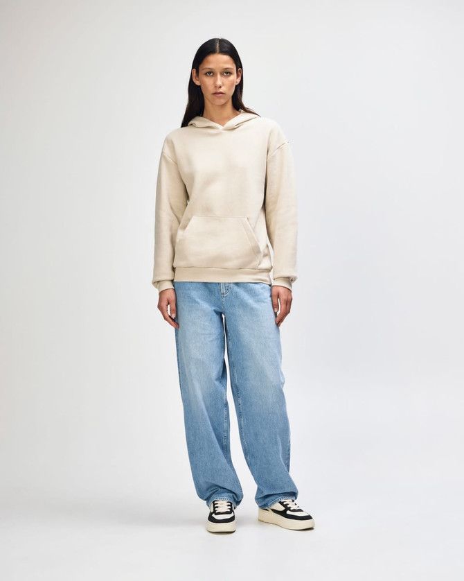 A unisex hooded sweatshirt in beige featuring a front pocket and a soft fleece material, worn with light blue jeans.
