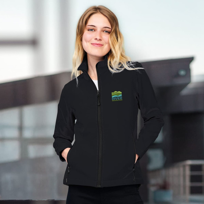A black women's softshell jacket with a zip front and a logo on the chest, worn by a model indoors.