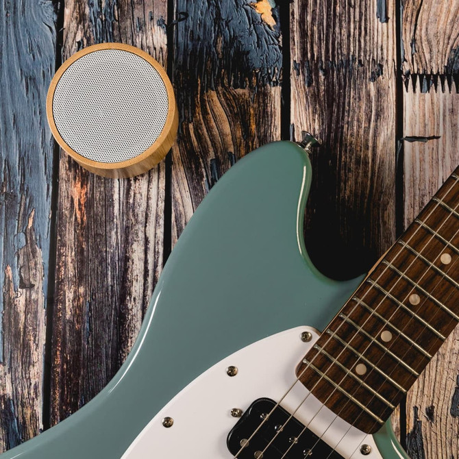 A light green guitar rests beside a round bamboo Bluetooth speaker on a rustic wooden surface.