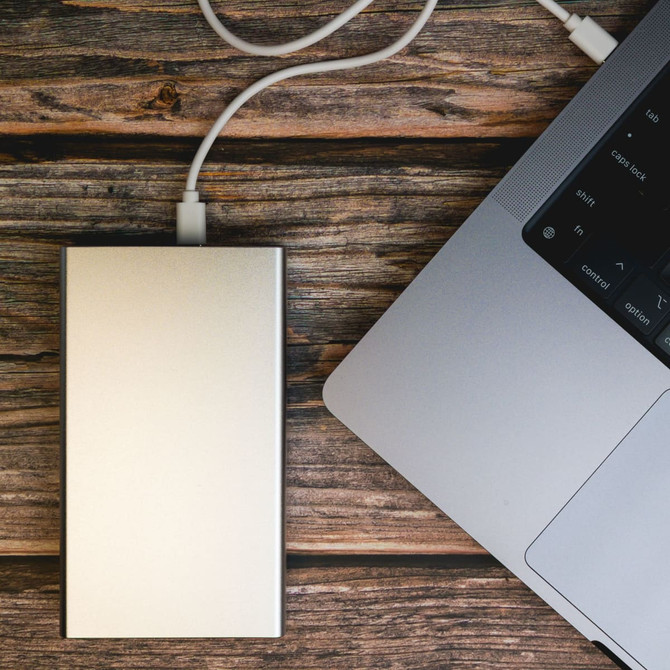A power bank, labeled Ortega, with a sleek silver design, connected to a laptop via a charging cable.