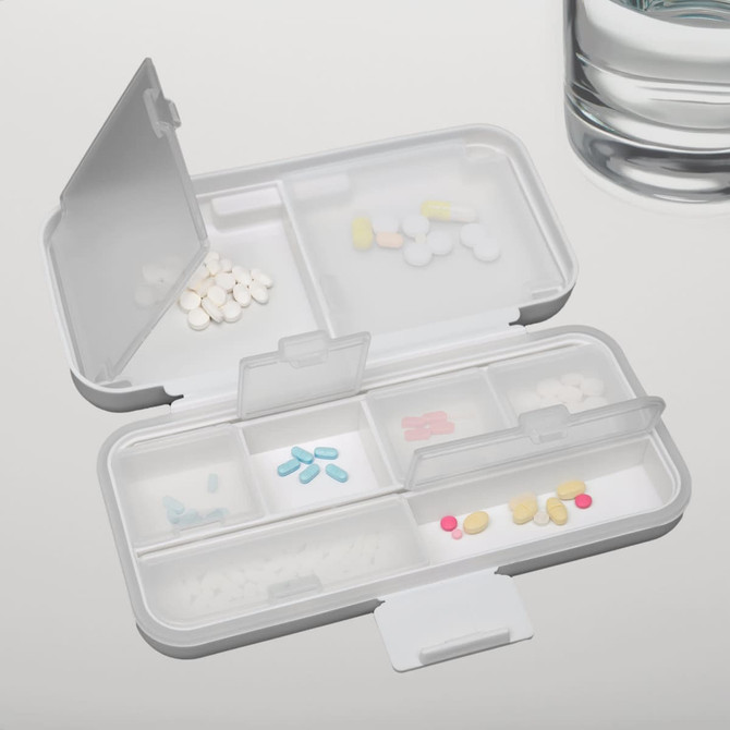 A compact white pill organizer with multiple compartments, filled with various colored tablets, beside a glass of water.