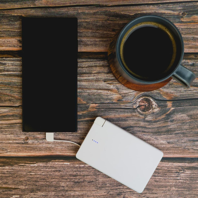 A white power bank is placed next to a black phone and a dark coffee mug on a wooden surface.