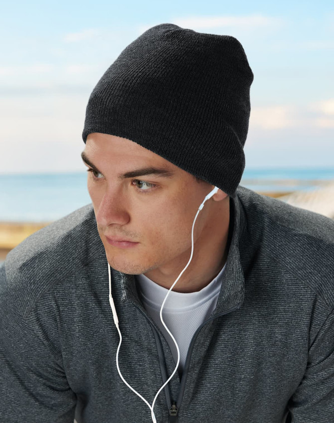 Acrylic knitted marl beanie in dark grey worn by a model with earbuds, set against a coastal background.
