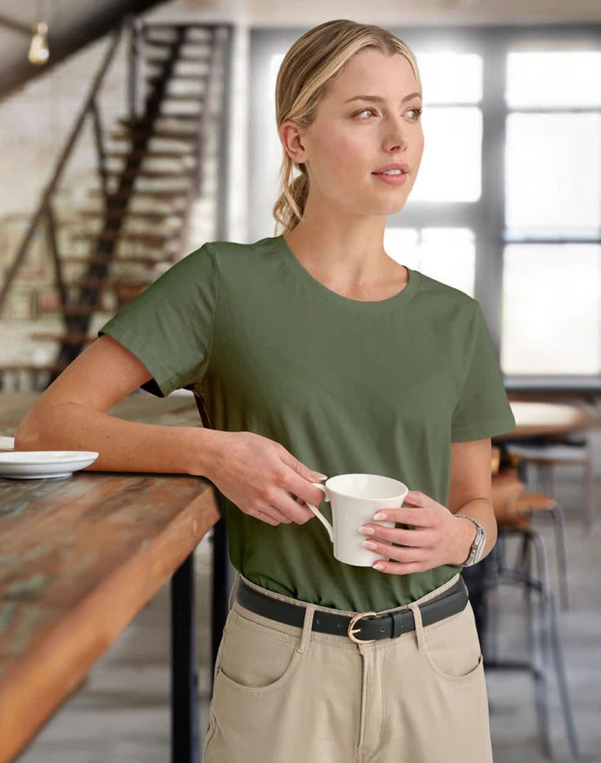 A woman wearing a green premium cotton tee, holding a white cup while standing indoors in a casual setting.