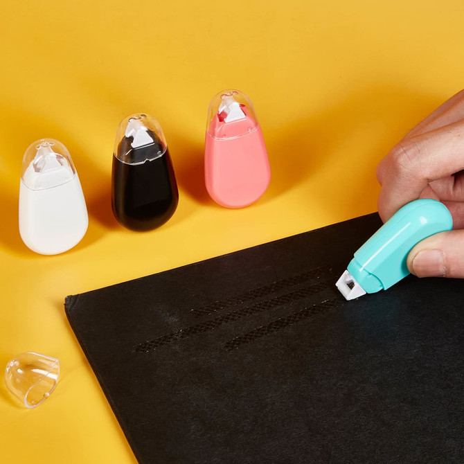 A hand using a turquoise Bayla Glue Tape on a black surface, with white, black, and pink glue tape holders in the background.