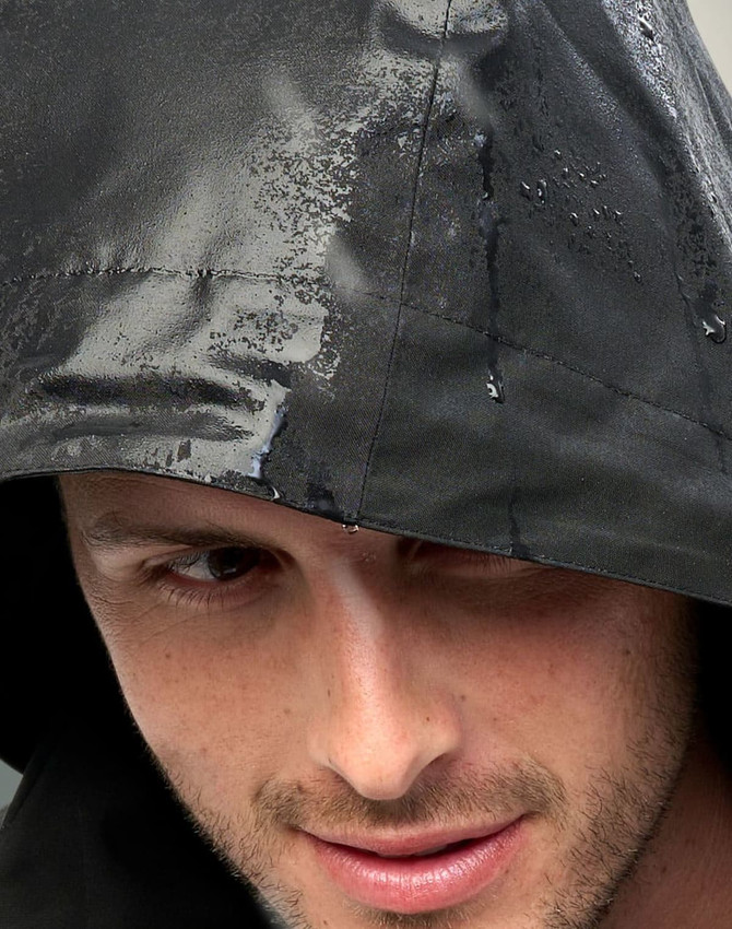 A close-up of a man wearing a black quilted waterproof jacket with a hood, featuring water droplets on the fabric.