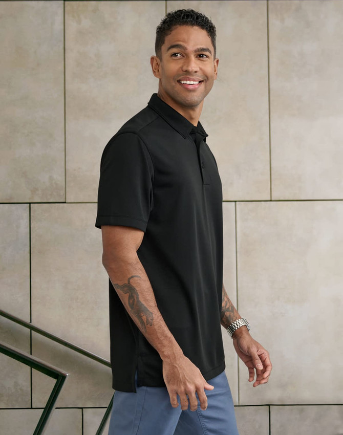A man wearing a black short-sleeve polo shirt stands against a textured wall, showcasing a sleek design and fit.