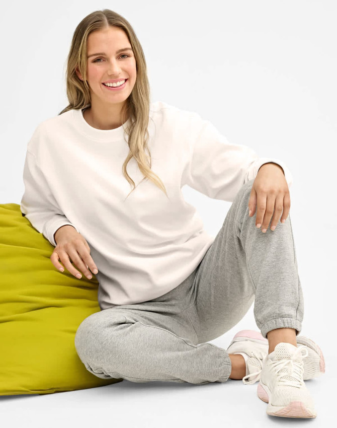 A woman sitting on a green bean bag, wearing grey sweatpants and a white sweatshirt, smiling at the camera.