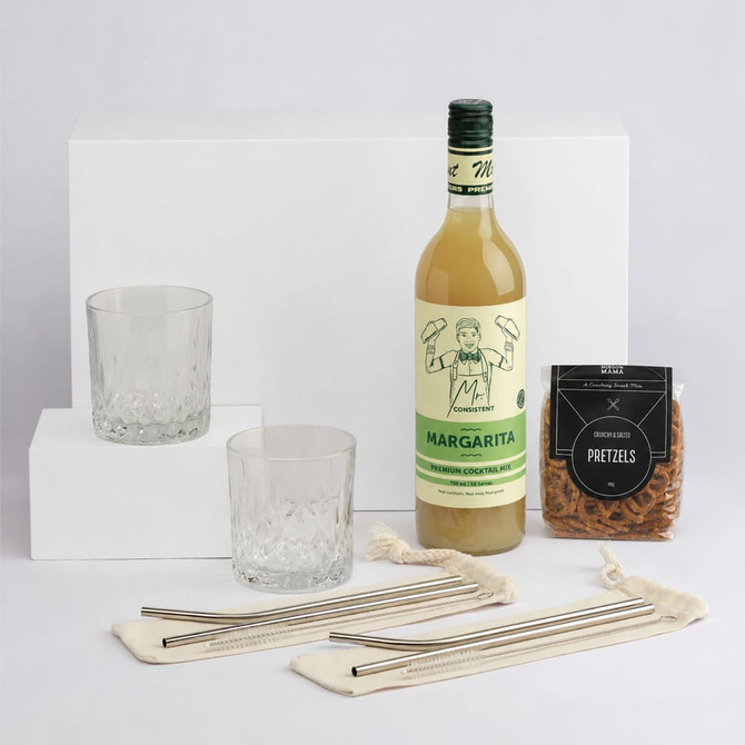 A green bottle of Margarita, two crystal glasses, a packet of pretzels, and metal straws on a white backdrop.