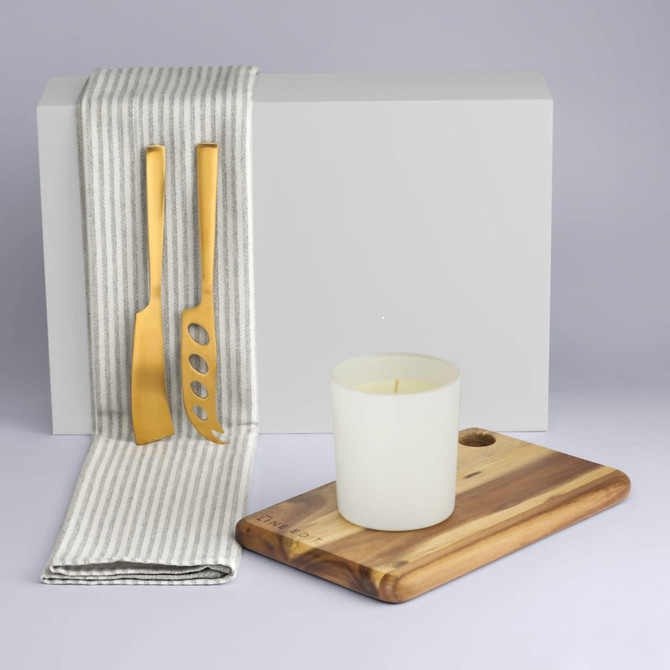 A gold cheese knife and slicer set on a grey and white striped cloth, alongside a white candle and wooden board.