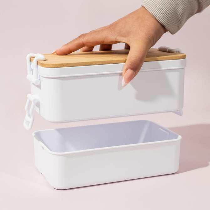 A white double-layer lunch box with a bamboo lid, being held by a hand. It features a branding logo.