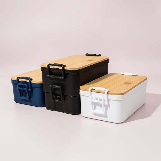Three lunch boxes in navy blue, black, and white with bamboo lids and handles, displaying a sleek, modern design.