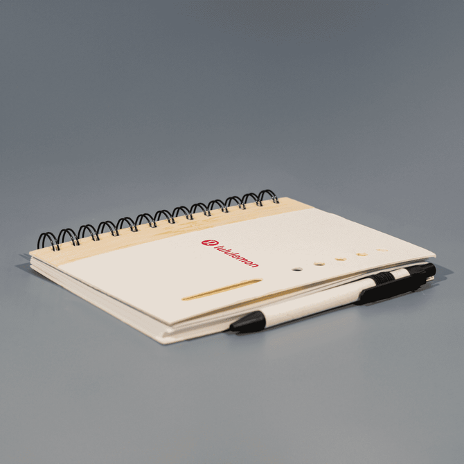 A spiral-bound notebook in cream with a black pen resting on it, featuring a logo on the cover.