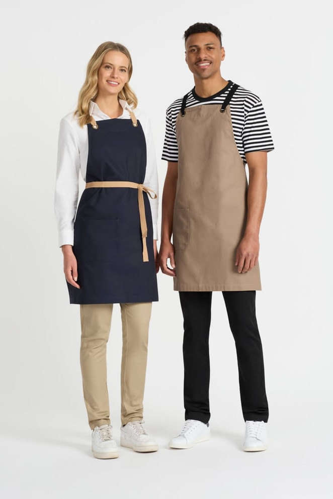 A navy bib apron on a woman and a beige bib apron on a man, both with shoulder straps, standing together.