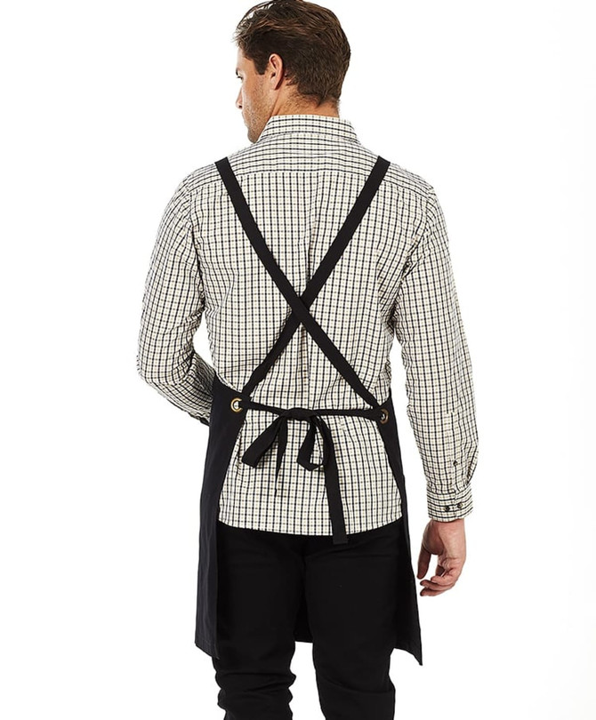 A black bib apron with crossed straps is displayed on a person wearing a checked shirt. The apron features a logo.