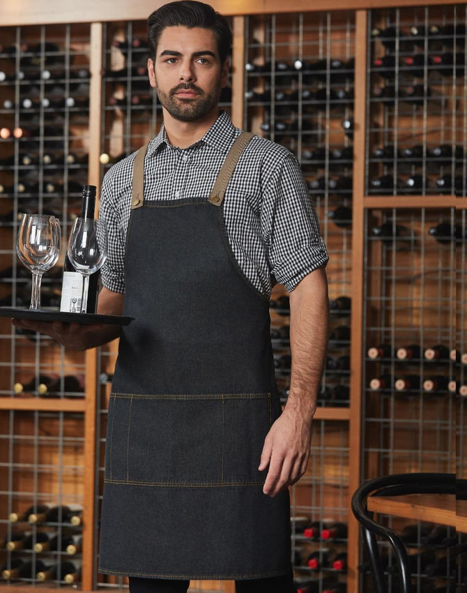 A denim bib apron in dark blue, worn by a man holding a tray with glasses and a bottle in a wine cellar setting.